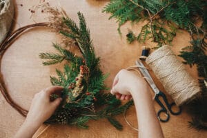 Hands holding fir branches and pine cones, berries, thread, scissors on wooden table. Making rustic christmas wreath flat lay. Details for christmas wreath workshop